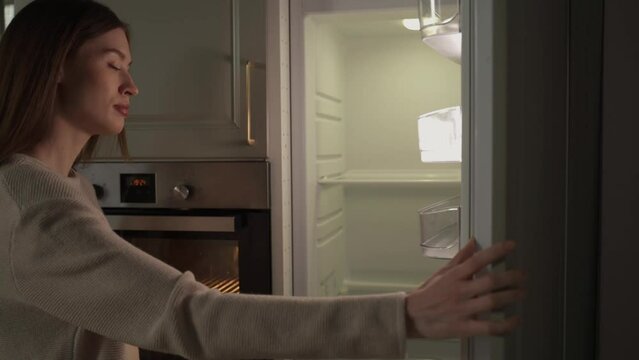 A young woman wearing a casual sweater stands pensively before an open, empty refrigerator in a dimly lit, modern kitchen environment, possibly considering her meal options or the need to go grocery