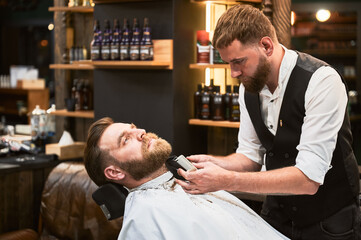 Professional barber using electric razor to shave beard. Regular client at an appointment in barbershop. Confident male customer getting his beard done.
