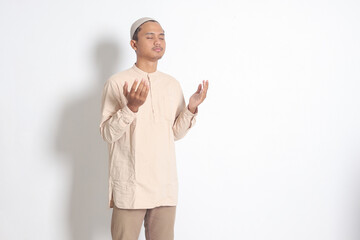 Portrait of religious Asian muslim man in koko shirt with skullcap praying earnestly with his hands...