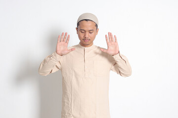 Portrait of religious Asian muslim man in koko shirt with skullcap praying with hands raised, doing...