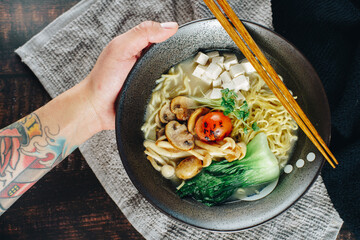A hand holding a bowl of noodles with mushrooms and tomatoes
