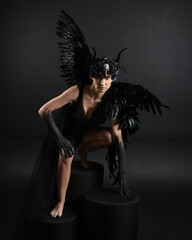 Full length portrait of model wearing gothic horned headdress with halloween black dress, fantasy angel feather wings. Crouching pose. Isolated dark studio background, cinematic shadow silhouettes 