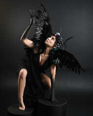 Full length portrait of model wearing gothic horned headdress with halloween black dress, fantasy angel feather wings. Crouching pose. Isolated dark studio background, cinematic shadow silhouettes 
