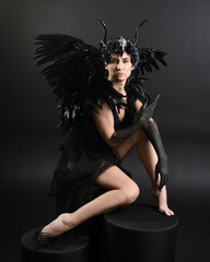 Full length portrait of model wearing gothic horned headdress with halloween black dress, fantasy angel feather wings. Crouching pose. Isolated dark studio background, cinematic shadow silhouettes 