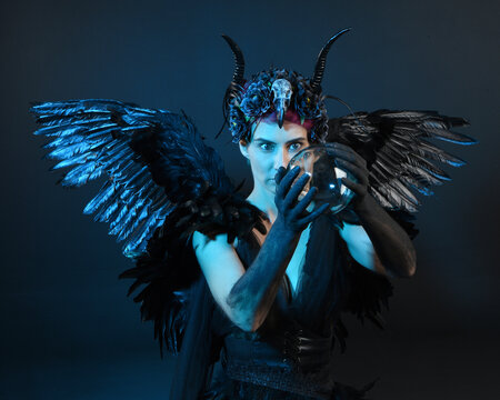 Close up portrait of female model wearing gothic horned headdress, halloween black dress, fantasy angel feather wings. Holding mystical crystal ball. dark studio background cinematic shadow silhouette - Powered by Adobe