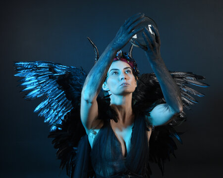 Close up portrait of female model wearing gothic horned headdress, halloween black dress, fantasy angel feather wings. Holding mystical crystal ball. dark studio background cinematic shadow silhouette