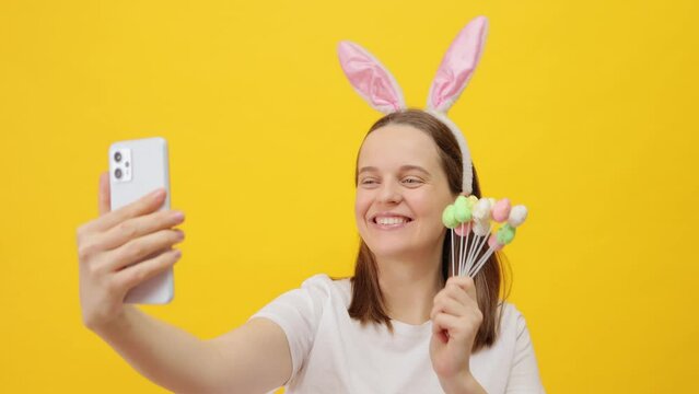 Satisfied delighted beautiful Caucasian woman wearing rabbit ears headband posing isolated over yellow background holding cake pops and making selfie on smartphone during Easter celebration