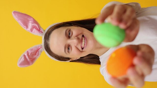 Vertical video. Happy satisfied Caucasian woman wearing rabbit ears headband showing multicolored eggs rejoicing happy Easter celebration posing isolated over yellow background