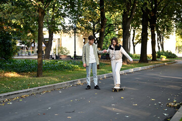 A young man teaches his girlfriend to skateboard on an alley in the park