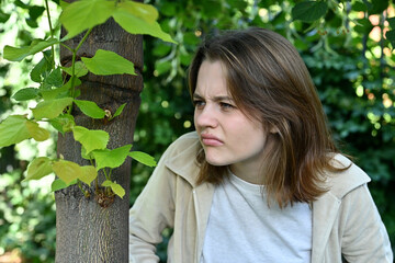 young woman stands behind a tree and hides and looks at something with disgust