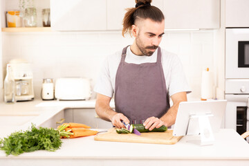 long-haired man cooking at home looking at tablet, chopping vegetables