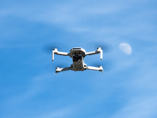 Bottom view of a small white drone with camera flying with its propellers out of focus due to speed and a blue sky with clouds and the Moon satellite in the background