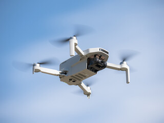 Bottom view of a small white drone with camera flying with its propellers out of focus due to speed flying through the air and a blue sky with clouds in the background