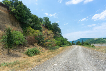 Gravel road through a river canyon. A hillock with trees and bushes on the right side. Bright day, clear blue sky with clouds. Georgia.