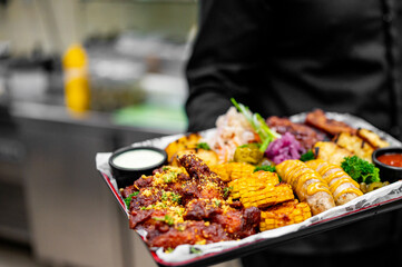 A person holds a tray of assorted grilled food, including ribs, corn, and sausages, ready to be served. The background is blurred, resembling a kitchen or serving area