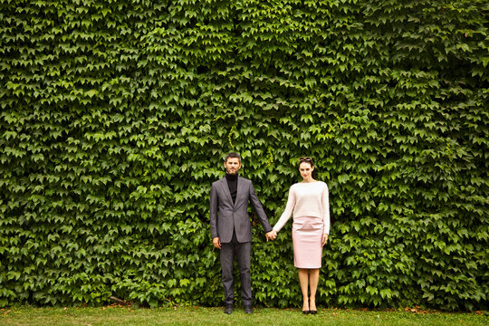 Couple Holding Hands Standing in front of Green Leafy Hedge
