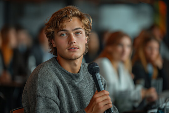 A young male with blond hair and blue eyes wearing a gray sweater holding a microphone