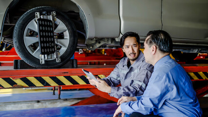 Asian automotive mechanic use tablet explain car condition to client in garage. Vehicle service manager male point at vehicle part to customer man for maintenance and repair car engine, auto service