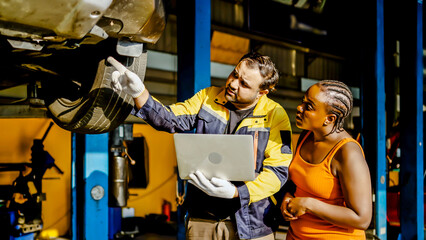 Asian automotive mechanic use laptop explain car condition to black woman client in garage. Vehicle service manager male point at vehicle part to customer man for maintenance and repair car engine