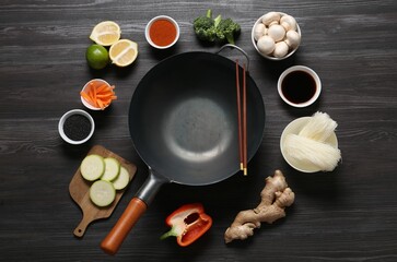 Empty iron wok and chopsticks surrounded by ingredients on dark grey wooden table, flat lay