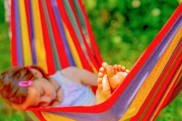 Gardinen Young beautiful girl sleeping in a hammock with bare feet, relaxing and enjoying a lovely sunny summer day. Green vegetation in background. Happy childhood concept © zwiebackesser