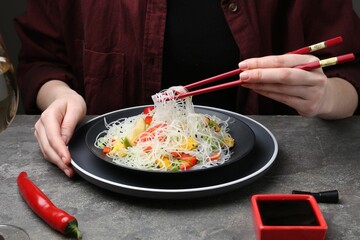 Stir-fry. Woman with chopsticks eating tasty rice noodles with meat and vegetables at grey textured table, closeup