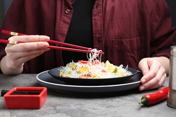 Stir-fry. Woman with chopsticks eating tasty rice noodles with meat and vegetables at grey textured table, closeup