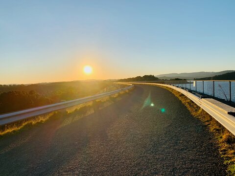 A road with a railing alongside a beautiful forest field and a serene waterscape during sunset