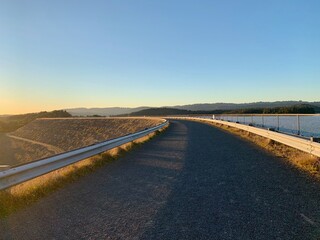 A road with a railing alongside a beautiful forest field and a serene waterscape during sunset - Powered by Adobe