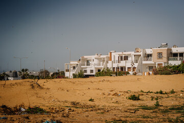 Agadir, Morocco - February 25, 2024 - Row of white houses behind a sandy open space with litter and sparse vegetation under a blue sky...