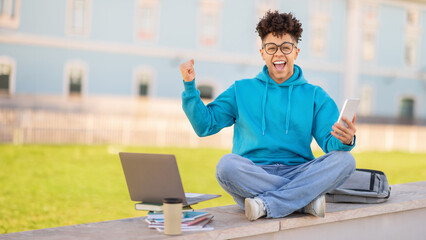 Joyful student guy with smartphone and laptop shaking fists outside