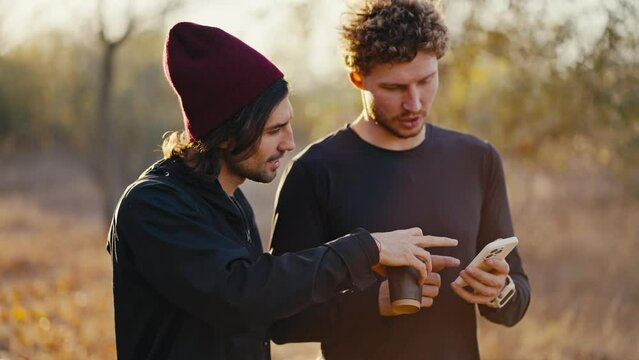 Close-up shot of two brunette athlete friends in a black sports uniform walking and talking while looking at something on the smartphone screen during their walk along an earthly path in a sunny