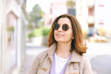 Outdoor portrait of happy middle age woman walking outside, wearing beige quilted jacket and sunglasses, nice sunny day