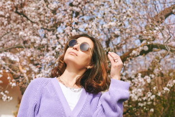 Outdoor fashion portrait of beautiful woman posing next to blooming spring tree, wearing purple knitted cardigan - 752174442