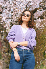 Outdoor fashion portrait of beautiful woman posing next to blooming spring tree, wearing purple knitted cardigan - 752174443