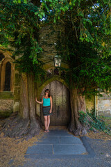 A young lady stands by a fairy-tale wooden door flanked by two old yew trees