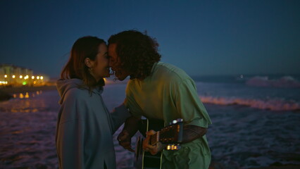 Fototapeta premium Enamoured man playing guitar at night sea beach closeup. Guy kissing girlfriend