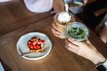 Top of hands of teenage girls holding glass of ice coffee and ice match green tea together  with blurred strawberry cheesecake at cafe.