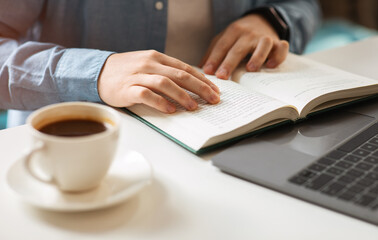 Close-up of a person's hands reading a book with a cup of coffee on the side and a laptop in the background