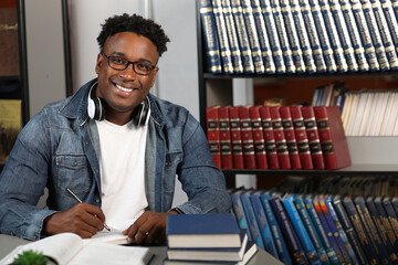 Young modern African businessman smiles, looks at the camera, sits at a desk in his office, writes...