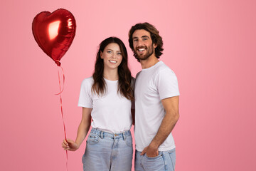 Cheerful young couple standing close together, with the woman holding a red heart-shaped balloon