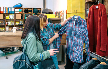 Female client looking plaid shirt while receives personalized support from friendly shop assistant. Blonde woman employee showing clothes to customer in vintage store. Small business concept.