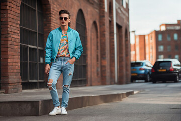 Fototapeta na wymiar Stylish young man wearing torn jeans and retro sunglasses, posing on city street. 90s fashion style and lifestyle