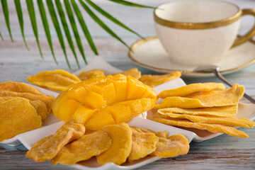 ripe juicy chopped mango is laid out on white platter along with dry plastics of fruit. On wooden table, tea pair on background of palm leaf and tropical mango on porcelain plate of irregular shape