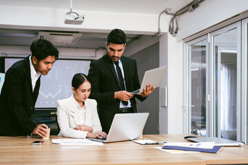 A cheerful and confident Asian businesswoman stands in the back of her office colleagues, presenting bar charts data from a monitor and projector screen. Asian businesswoman leader role at the meeting