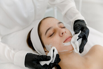 A cosmetologist removing a mask from the skin of a teenage girl’s face by cleaning wipes. Teen...