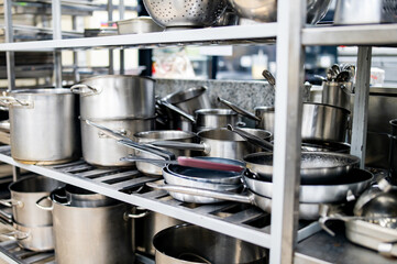 photo showcases various sizes of stainless steel cooking utensils neatly arranged on a metal shelving unit in a professional kitchen environment. Large pots, frying pans, and a colander are visible. - obrazy, fototapety, plakaty