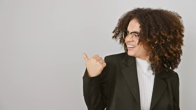 Cheerful hispanic woman with curly hair smiling and pointing to the side with thumb up, standing against isolated white background