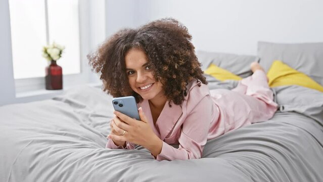 Joyful, curly hair hispanic woman lying on bed, happily pointing to one side with a confident smile. engaged in texting on smartphone in bedroom scene.