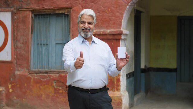 Old age man dressed in formal wear feeling happy after giving his vote in the elections - responsible citizen  India. Grey hair businessman showing his voter identification card with a thumbs up ac...